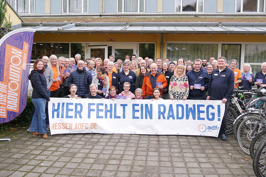 Gruppenfoto 2 Die Gruppe der ADFC-Aktiven steht vor dem Eingang und hält ein Banner mit der Aufschrift „Hier fehlt ein Radweg“ in die Höhe
