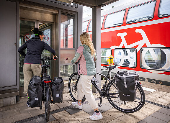 Sollte unkompliziert sein: Fahrradmitnahme in der Bahn. Zwei Frauen schieben zwei Fahrräder in einen Aufzug am Bahnhof. Im Hinterung ein Regionalzug mit Fahrradsymbol.